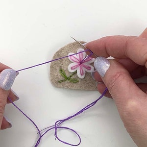 May include: Close-up of a needle and thread being used to embroider a felt patch. The patch features a white and pink flower with green leaves. The thread is purple, and the patch is beige.