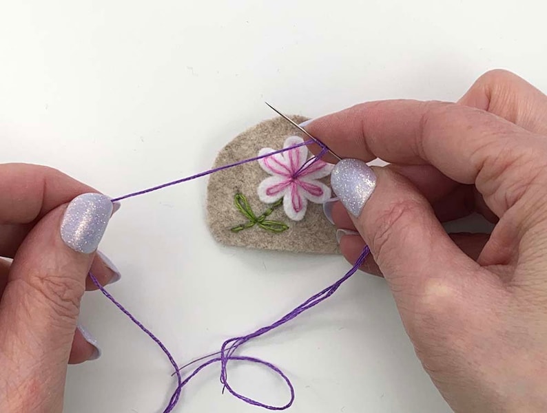 May include: Close-up of a needle and thread being used to embroider a felt patch. The patch features a white and pink flower with green leaves. The thread is purple, and the patch is beige.
