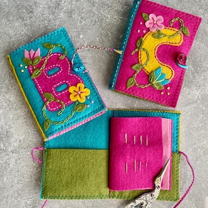 Könnte beinhalten: Handgefertigtes Nähset aus Filz in Türkis, Pink und Grün. Das Set zeigt gestickte Blumendesigns und den Buchstaben "B" auf dem Deckel. Enthält einen Nadelhalter, eine Schere und einen Knopfverschluss. Das Set ist etwa 10 cm breit und 13 cm hoch.