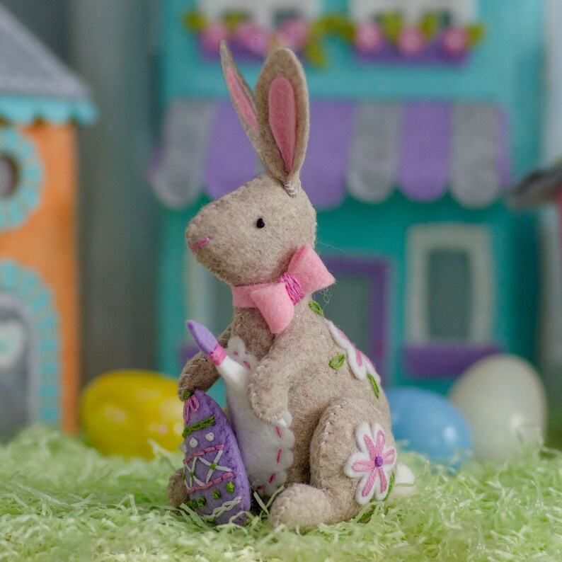 May include: A tan felt bunny figurine with a pink bow, holding a purple Easter egg. The bunny has pink-lined ears and is decorated with embroidered flowers. The scene includes colorful toy houses and Easter eggs on green paper grass.
