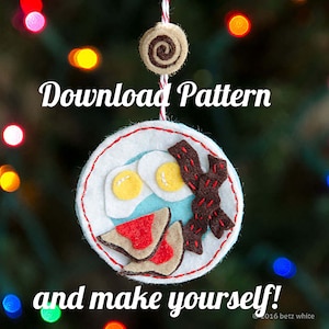Może przedstawiać: Ozdoba filcowa w kształcie talerza z motywem śniadaniowym. Talerz zawiera dwa jajka sadzone, bekon i kromkę tostów. Tekst "Download Pattern and make yourself!" znajduje się pod ozdobą.