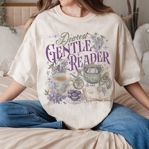 Könnte beinhalten: Cremefarbenes T-Shirt mit dem Text "Dearest Gentle Reader". Die Grafik zeigt eine Teetasse, eine Kutsche, ein offenes Buch und florale Elemente in Lila und Grün. Das Shirt hat eine lockere Passform.