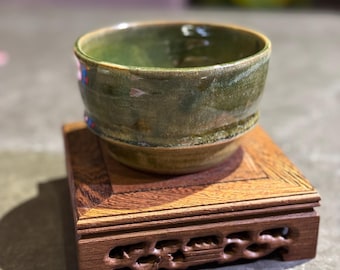May include: A green ceramic teacup with a brown rim sits on a dark wood stand with intricate carvings. The stand has a square top and a decorative base.