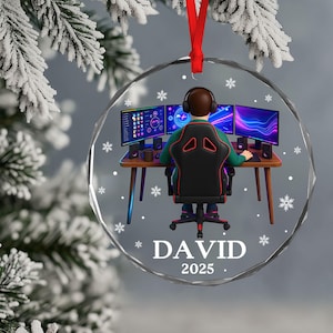 Puede incluir: Un adorno navideño redondo y transparente con una cinta roja. El adorno presenta a un jugador en un escritorio con tres monitores, auriculares y el nombre "DAVID" y el año "2025". Copos de nieve están dispersos.