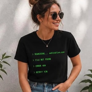 May include: Black t-shirt with green text that reads "SEARCHING... motivation.exe", "FILE NOT FOUND", "ERROR 404", and "RETRY? Y/N". The shirt is worn by a person wearing sunglasses and blue jeans.