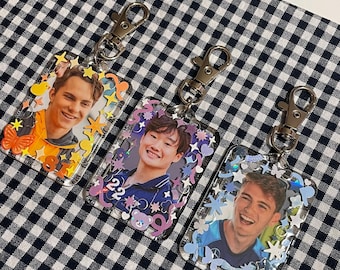 Può includere: Tre portachiavi in plastica trasparente con foto diverse di giovani uomini all'interno. Ogni portachiavi ha un anello in metallo argentato e una chiusura in metallo argentato. I portachiavi sono decorati con glitter colorati e piccole stelle, cuori e altre forme in plastica.