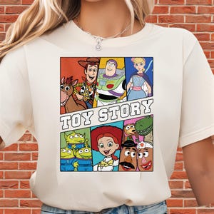 May include: Cream-colored t-shirt featuring a colorful graphic of characters from the animated film "Toy Story." The design includes Woody, Buzz Lightyear, Bo Peep, and other characters, with the words "TOY STORY" in bold letters.