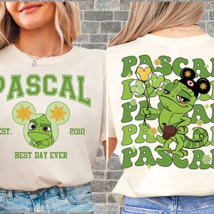 May include: Cream-colored t-shirt featuring a green graphic of Pascal the chameleon from the movie Tangled. The front design includes the word "PASCAL" and a Mickey Mouse-shaped head with Pascal's face. The back repeats "PASCAL" with Pascal holding flowers.