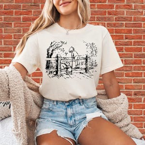 Puede incluir: Camiseta color crema con una ilustración en blanco y negro de una escena de cuento infantil clásico. El gráfico representa a un niño y un oso interactuando cerca de una valla de madera. La camiseta se combina con pantalones cortos vaqueros.