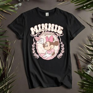Puede incluir: Camiseta negra con un dise&ntilde;o retro de Minnie Mouse. El dise&ntilde;o incluye el texto "MINNIE Mouse" en una fuente de burbujas, con Minnie sosteniendo una taza de t&eacute;, enmarcada por un &oacute;valo rosa y hojas de laurel. La camiseta se muestra sobre un fondo neutro.