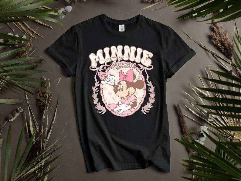 Puede incluir: Camiseta negra con un dise&ntilde;o retro de Minnie Mouse. El dise&ntilde;o incluye el texto "MINNIE Mouse" en una fuente de burbujas, con Minnie sosteniendo una taza de t&eacute;, enmarcada por un &oacute;valo rosa y hojas de laurel. La camiseta se muestra sobre un fondo neutro.