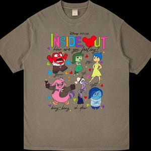 May include: A tan Inside Out t-shirt featuring the Disney Pixar logo and characters. The graphic includes the words "INSIDE OUT" and "how are you feeling?" with images of the characters Joy, Sadness, Anger, Fear, Disgust, and Bing Bong.