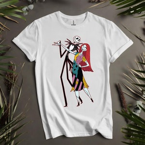 May include: White t-shirt with a cartoon illustration of Jack Skellington and Sally from The Nightmare Before Christmas. Jack, a skeletal figure, and Sally, a ragdoll with red hair and a patchwork dress, are depicted. The design is centrally placed on the shirt.