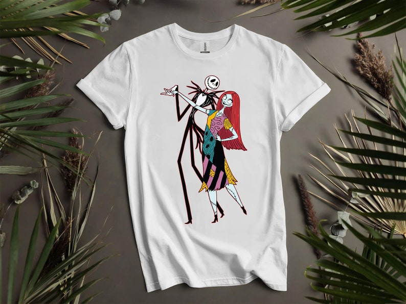 May include: White t-shirt with a cartoon illustration of Jack Skellington and Sally from The Nightmare Before Christmas. Jack, a skeletal figure, and Sally, a ragdoll with red hair and a patchwork dress, are depicted. The design is centrally placed on the shirt.