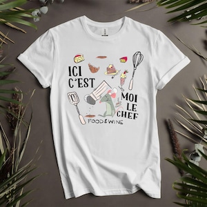 May include: White t-shirt with a cartoon mouse chef design, surrounded by food illustrations and the text "ICI C'EST MOI LE CHEF FOOD & WINE". The design includes a whisk, spatula, and other cooking elements.