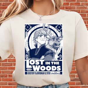 Puede incluir: Camiseta color crema con un gr&aacute;fico azul y blanco de personajes de la pel&iacute;cula Frozen II. El gr&aacute;fico incluye el texto "LOST IN THE WOODS" y los nombres "KRISTOFF BJORGMAN & SVEN".