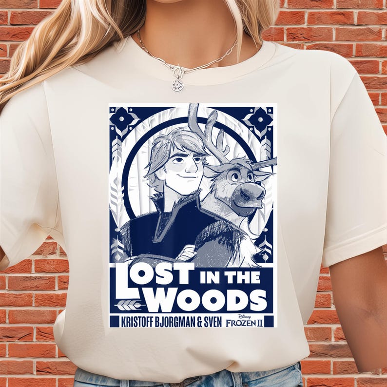Puede incluir: Camiseta color crema con un gr&aacute;fico azul y blanco de personajes de la pel&iacute;cula Frozen II. El gr&aacute;fico incluye el texto "LOST IN THE WOODS" y los nombres "KRISTOFF BJORGMAN & SVEN".