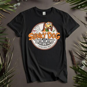Puede incluir: Camiseta negra con un gr&aacute;fico circular de Slinky Dog y el texto "Slinky Dog Dash" en naranja. El dise&ntilde;o incluye la frase "GO! GO! GO!" y "Established 2018". La camiseta se muestra sobre un fondo neutro con vegetaci&oacute;n.