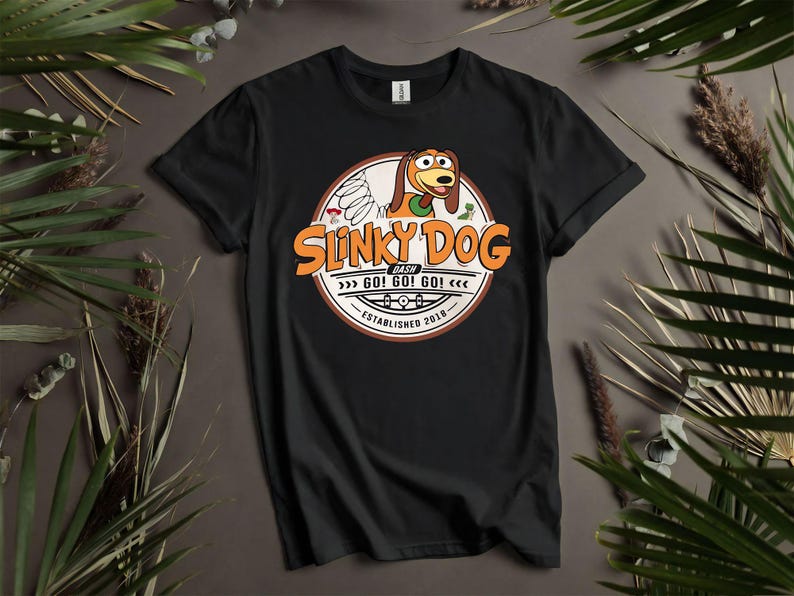 Puede incluir: Camiseta negra con un gr&aacute;fico circular de Slinky Dog y el texto "Slinky Dog Dash" en naranja. El dise&ntilde;o incluye la frase "GO! GO! GO!" y "Established 2018". La camiseta se muestra sobre un fondo neutro con vegetaci&oacute;n.