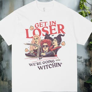 Puede incluir: Camiseta blanca con un gráfico que presenta a tres mujeres en un coche, un gato negro y el texto "GET IN LOSER WE'RE GOING WITCHIN'". El diseño incluye calabazas y murciélagos de dibujos animados, perfecto para Halloween.