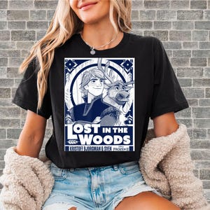 Puede incluir: Camiseta negra con un dise&ntilde;o gr&aacute;fico de la pel&iacute;cula de Disney Frozen II. El dise&ntilde;o incluye el texto "LOST IN THE WOODS" con im&aacute;genes de Kristoff, Sven y elementos decorativos en blanco y azul marino.