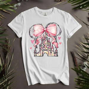 May include: White t-shirt featuring a pink bow and a castle design. The castle is pink and yellow with a pink ribbon. The design includes pink hearts and Mickey Mouse silhouettes. The shirt is a short-sleeved crew neck.