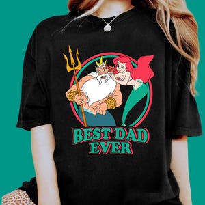 May include: Black t-shirt featuring a colorful graphic of King Triton holding a trident, with Ariel and the text "BEST DAD EVER" in green and white. The design is set within a circular frame.