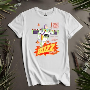 Puede incluir: Camiseta blanca con un gr&aacute;fico de Buzz Lightyear de Toy Story. El dise&ntilde;o incluye a Buzz en una pose de vuelo, las palabras "BUZZ LIGHTYEAR" en una explosi&oacute;n roja y amarilla, y el texto "DELUXE SPACE RANGER".