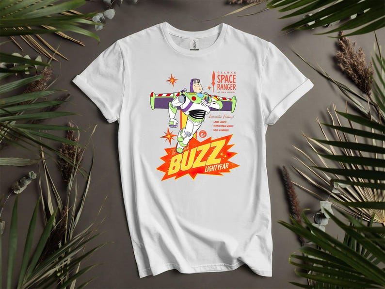 Puede incluir: Camiseta blanca con un gr&aacute;fico de Buzz Lightyear de Toy Story. El dise&ntilde;o incluye a Buzz en una pose de vuelo, las palabras "BUZZ LIGHTYEAR" en una explosi&oacute;n roja y amarilla, y el texto "DELUXE SPACE RANGER".