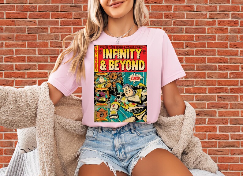 Puede incluir: Camiseta rosa claro con un dise&ntilde;o de c&oacute;mic. El gr&aacute;fico presenta las palabras "INFINITY & BEYOND" e ilustraciones de Buzz Lightyear y Zurg. La camiseta tiene mangas cortas y cuello redondo.