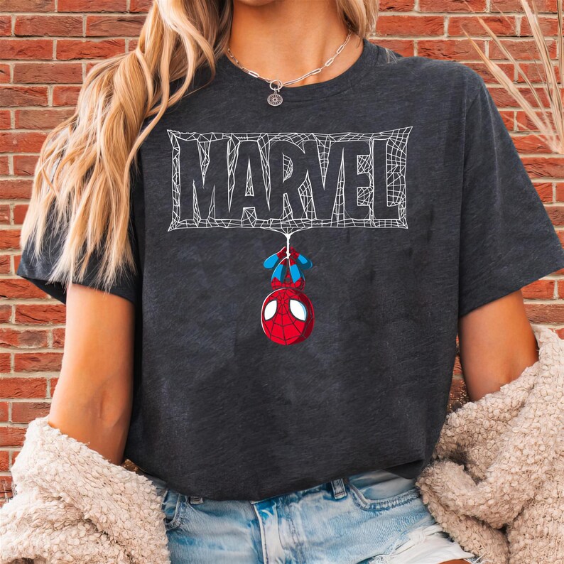 May include: Dark gray t-shirt featuring the word "MARVEL" in white, with a web design. A cartoon Spider-Man hangs below. The shirt is short-sleeved and has a crew neck.