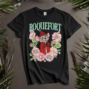 Peut inclure: T-shirt noir avec le mot "ROQUEFORT" en vert menthe au-dessus d'une souris de dessin anim&eacute; portant une cape et un chapeau rouges, entour&eacute;e de fleurs roses, vertes et blanches. Le t-shirt a un col rond.