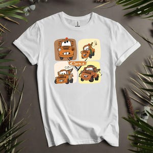 May include: White t-shirt featuring a colorful graphic of the character Mater from the animated movie Cars. The design includes four different images of Mater in square frames, with the word "Cars" in the center.