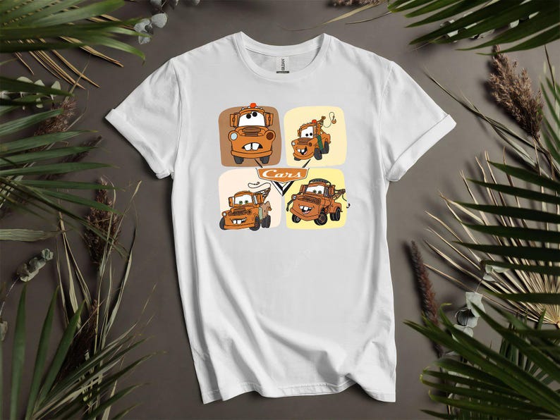 May include: White t-shirt featuring a colorful graphic of the character Mater from the animated movie Cars. The design includes four different images of Mater in square frames, with the word "Cars" in the center.