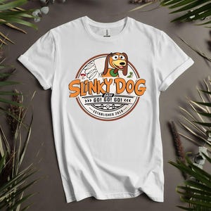 Puede incluir: Camiseta blanca con un dise&ntilde;o circular. El dise&ntilde;o presenta el texto "Slinky Dog Dash" en naranja y marr&oacute;n, una ilustraci&oacute;n de Slinky Dog y las palabras "GO! GO! GO!" y "Established 2018".