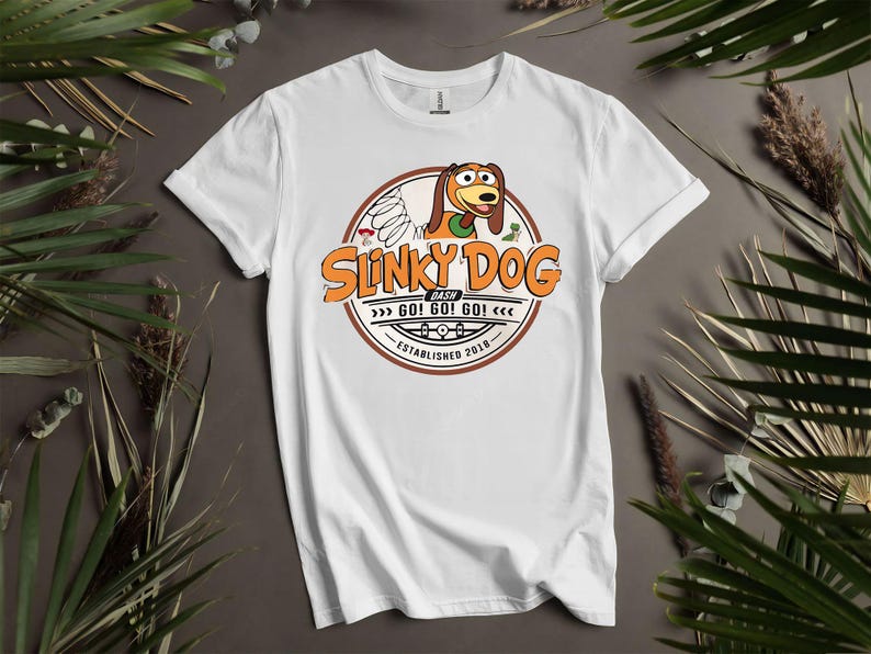 Puede incluir: Camiseta blanca con un dise&ntilde;o circular. El dise&ntilde;o presenta el texto "Slinky Dog Dash" en naranja y marr&oacute;n, una ilustraci&oacute;n de Slinky Dog y las palabras "GO! GO! GO!" y "Established 2018".