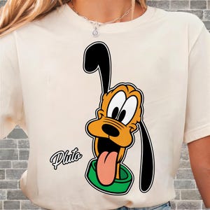 May include: Cream-colored t-shirt featuring a cartoon image of Pluto, a Disney character. Pluto is depicted with orange fur, black ears, and a green collar, with his tongue sticking out. The name "Pluto" is written in cursive below the character.