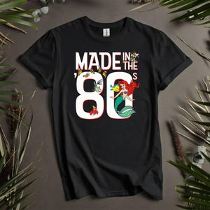 May include: Black t-shirt with the text "MADE IN THE 80'S" in white. The number 80 features a mermaid illustration with red hair and a green tail. Additional cartoon sea creatures are also present.