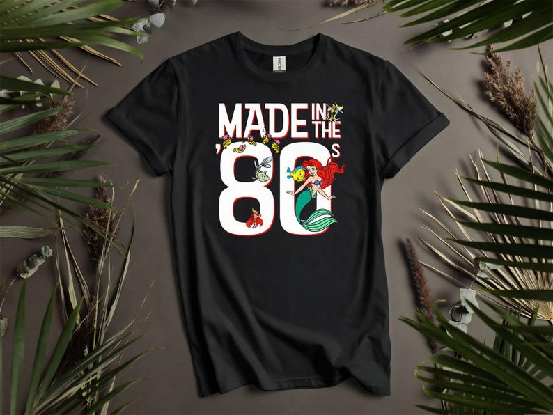 May include: Black t-shirt with the text "MADE IN THE 80'S" in white. The number 80 features a mermaid illustration with red hair and a green tail. Additional cartoon sea creatures are also present.