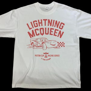 K&ouml;nnte beinhalten: Wei&szlig;es T-Shirt mit roter Grafik von Lightning McQueen, einem Rennwagen aus dem Film Cars. Das Design enth&auml;lt den Text "LIGHTNING McQUEEN" und "PISTON CUP RACING SERIES 5 TIME CHAMP".