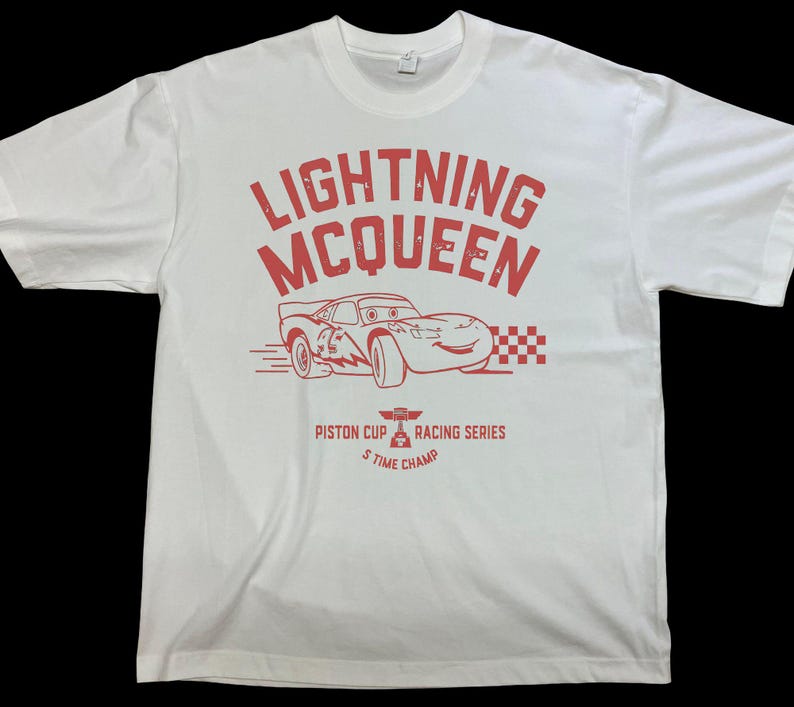 K&ouml;nnte beinhalten: Wei&szlig;es T-Shirt mit roter Grafik von Lightning McQueen, einem Rennwagen aus dem Film Cars. Das Design enth&auml;lt den Text "LIGHTNING McQUEEN" und "PISTON CUP RACING SERIES 5 TIME CHAMP".