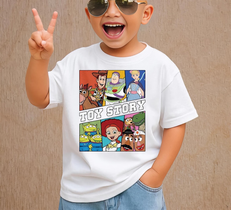 May include: White t-shirt featuring a colorful graphic of characters from the animated film "Toy Story." The design includes Woody, Buzz Lightyear, and other characters, with the words "TOY STORY" in bold letters.