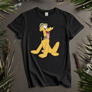 Könnte beinhalten: Schwarzes T-Shirt mit einem Cartoon-Bild von Pluto, dem Hund, der einen Safarihut trägt. Der gelbe Hund hat eine lange rosa Zunge, die herausragt. Das Shirt wird auf einem neutralen Hintergrund mit grünen Blättern präsentiert.