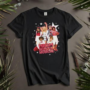 May include: Black t-shirt featuring a graphic of the High School Musical cast. The design includes the text "HIGH SCHOOL MUSICAL" in red and white, with star accents. The shirt is displayed on a neutral surface with greenery.
