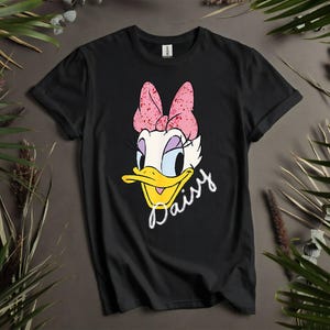 Peut inclure: T-shirt noir avec un dessin de Daisy Duck, personnage de dessin animé, orné d'un nœud rose et du nom "Daisy" en écriture blanche. Le t-shirt est de style col rond.