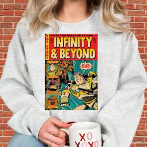 Puede incluir: Una sudadera gris claro con un gr&aacute;fico estilo c&oacute;mic. El gr&aacute;fico presenta las palabras "INFINITY & BEYOND" en rojo y amarillo, e im&aacute;genes de Buzz Lightyear y Zurg. El texto "TOY STORY" est&aacute; en la parte superior.