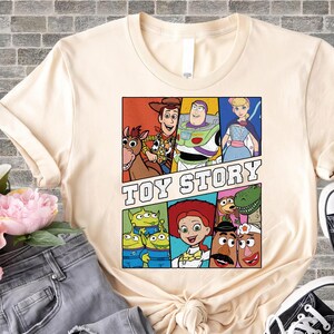 May include: Cream-colored t-shirt featuring a colorful graphic of characters from the animated film "Toy Story." The design includes Woody, Buzz Lightyear, Jessie, and other characters in a comic book style. The words "Toy Story" are printed in bold letters.