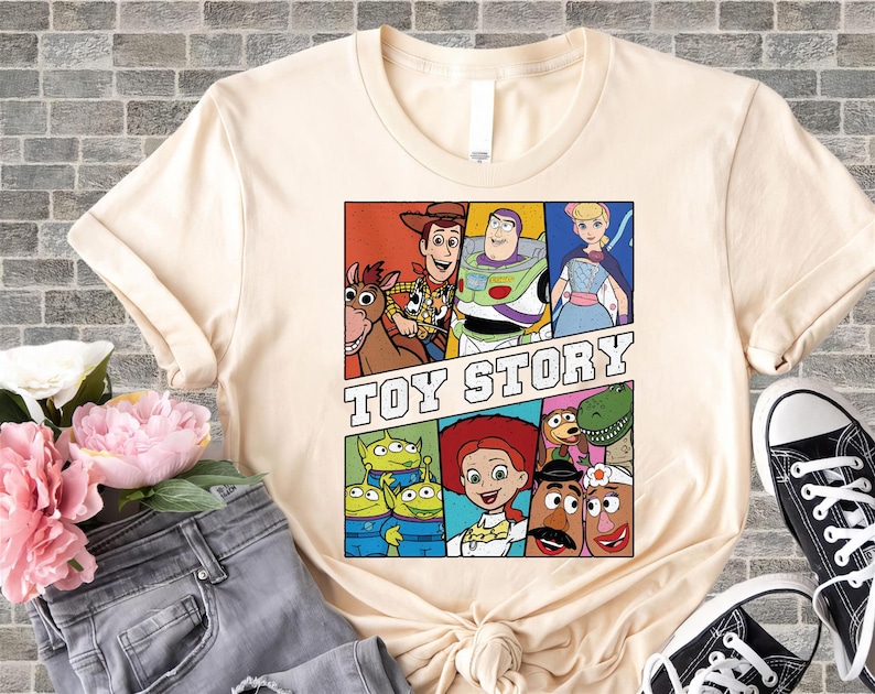 May include: Cream-colored t-shirt featuring a colorful graphic of characters from the animated film "Toy Story." The design includes Woody, Buzz Lightyear, Jessie, and other characters in a comic book style. The words "Toy Story" are printed in bold letters.