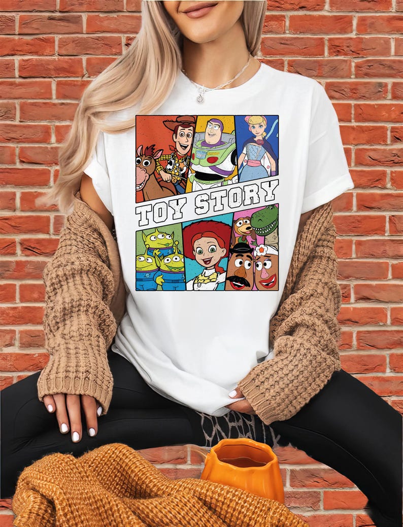 May include: White t-shirt featuring a colorful graphic of characters from the animated film "Toy Story." The design includes images of Woody, Buzz Lightyear, and other characters, with the words "TOY STORY" in bold letters. The shirt is a classic crew neck.