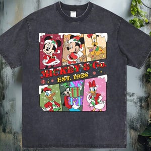 May include: Dark gray t-shirt featuring a colorful graphic of Mickey Mouse, Minnie Mouse, Donald Duck, Goofy, and Daisy Duck in Christmas attire. The text "Mickey & Co. EST. 1928" is also displayed. The shirt has a vintage, distressed look.